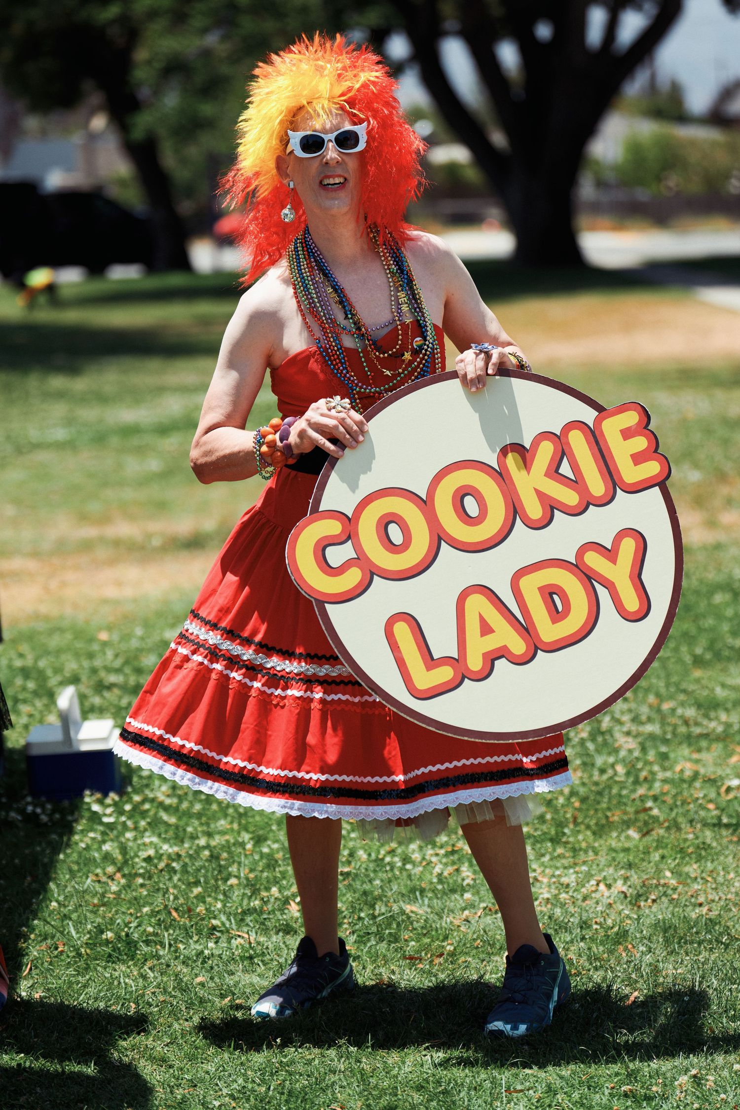 Colorfully dressed figure in red dress holding Cookie Lady sign wears orange wig and sunglasses while standing outdoors.