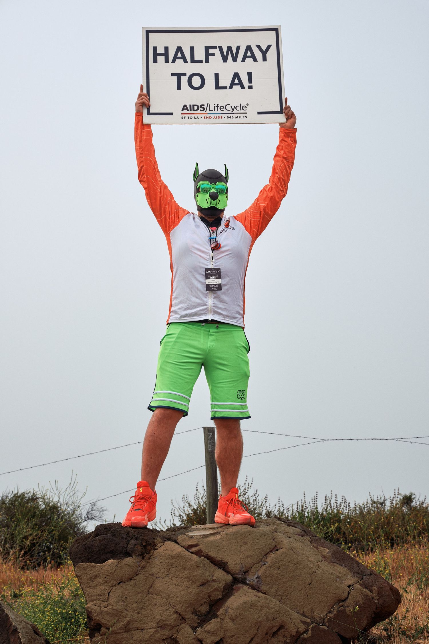 A cyclist in orange and green attire holds a 'Halfway to LA' sign while standing on a rock formation.