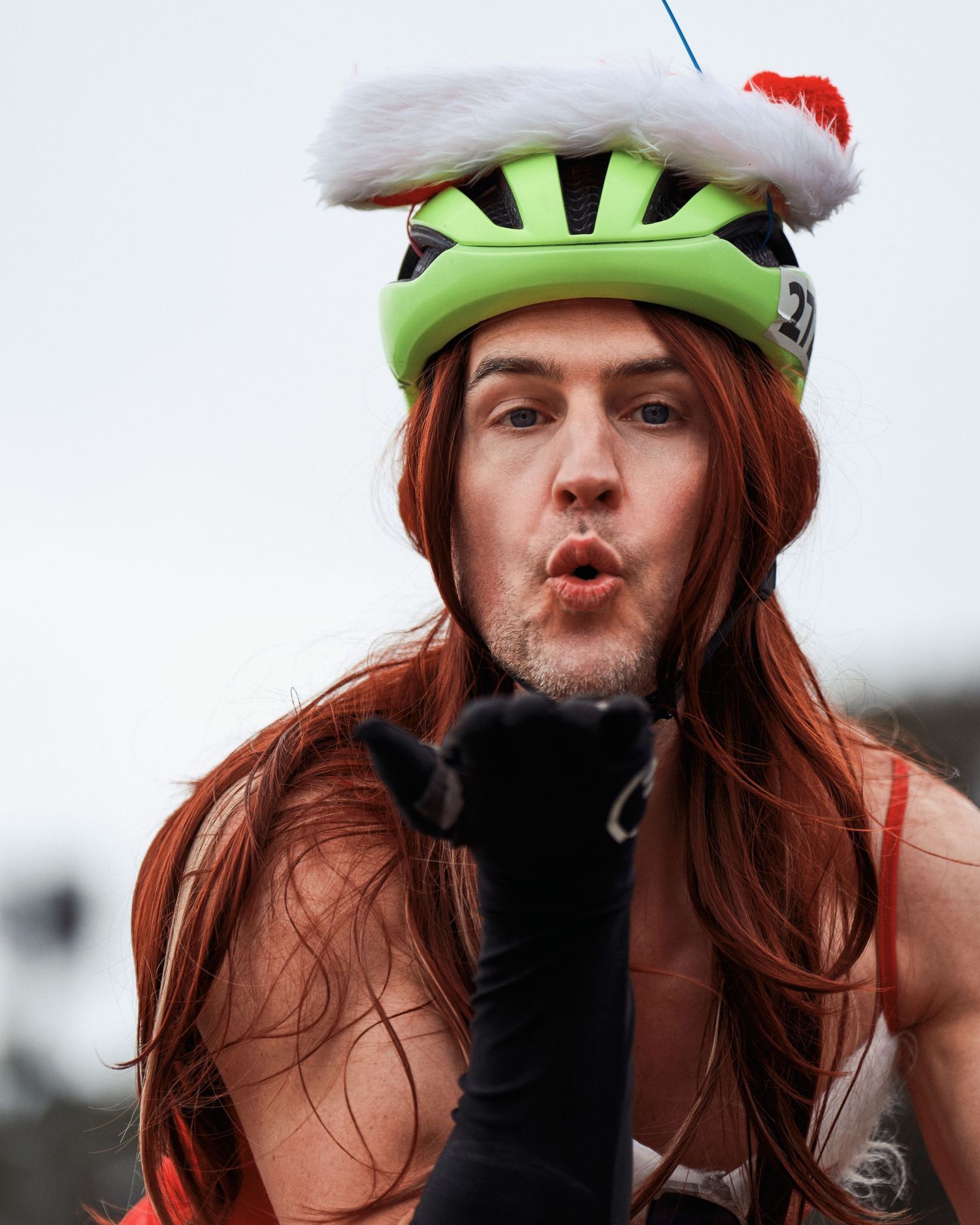 A cyclist wearing a green helmet with white fur trim and long hair makes a playful expression.