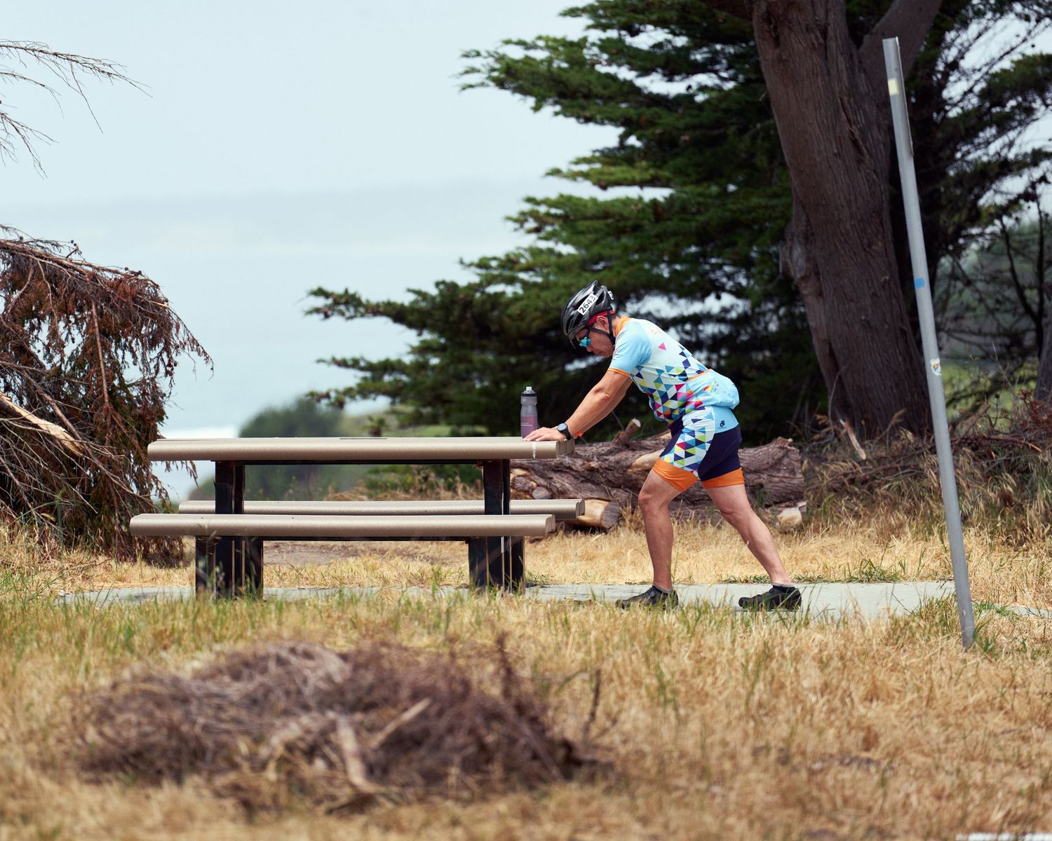 Cyclist stretches on wooden picnic bench surrounded by dry grass and cypress trees.