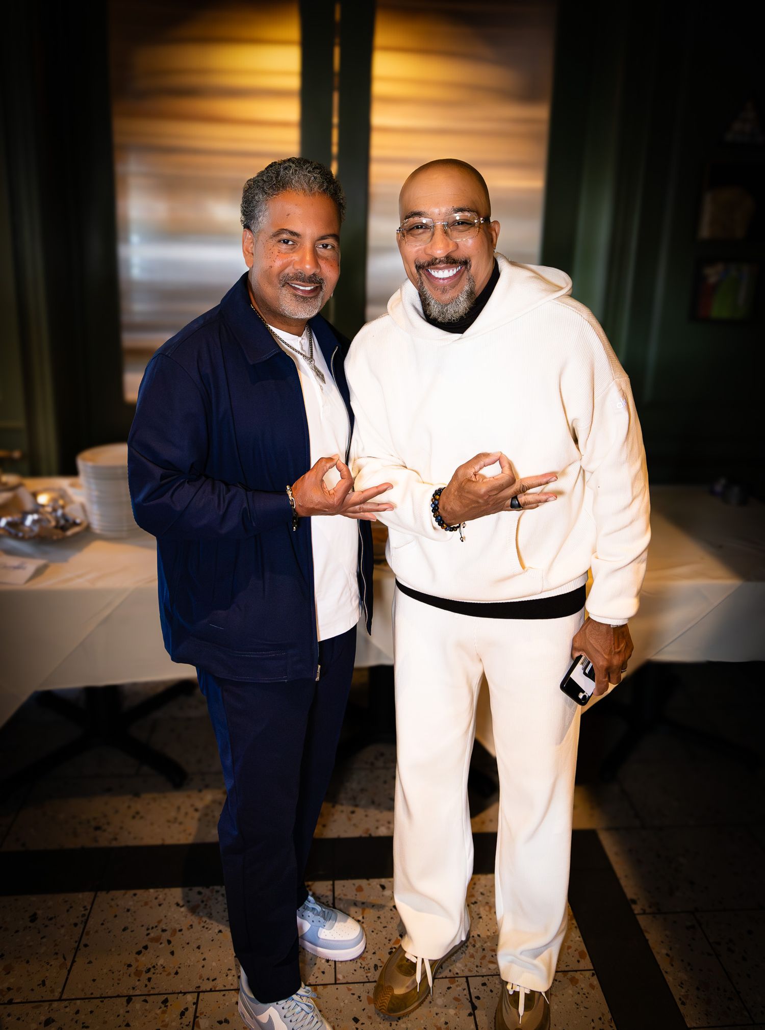 Two men stand together indoors, one wearing a navy jacket and the other in all white attire, both smiling and gesturing.