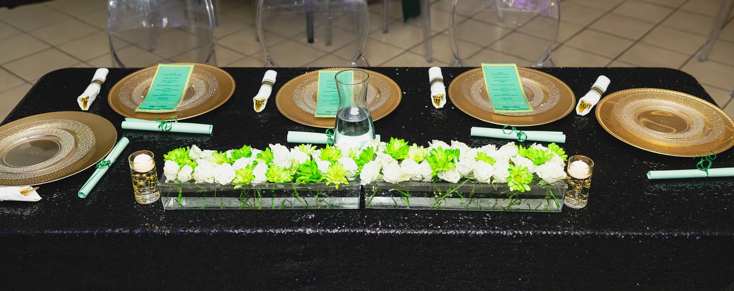 Elegant table setting with gold chargers, teal napkins, white floral centerpiece, and black tablecloth for a formal event.