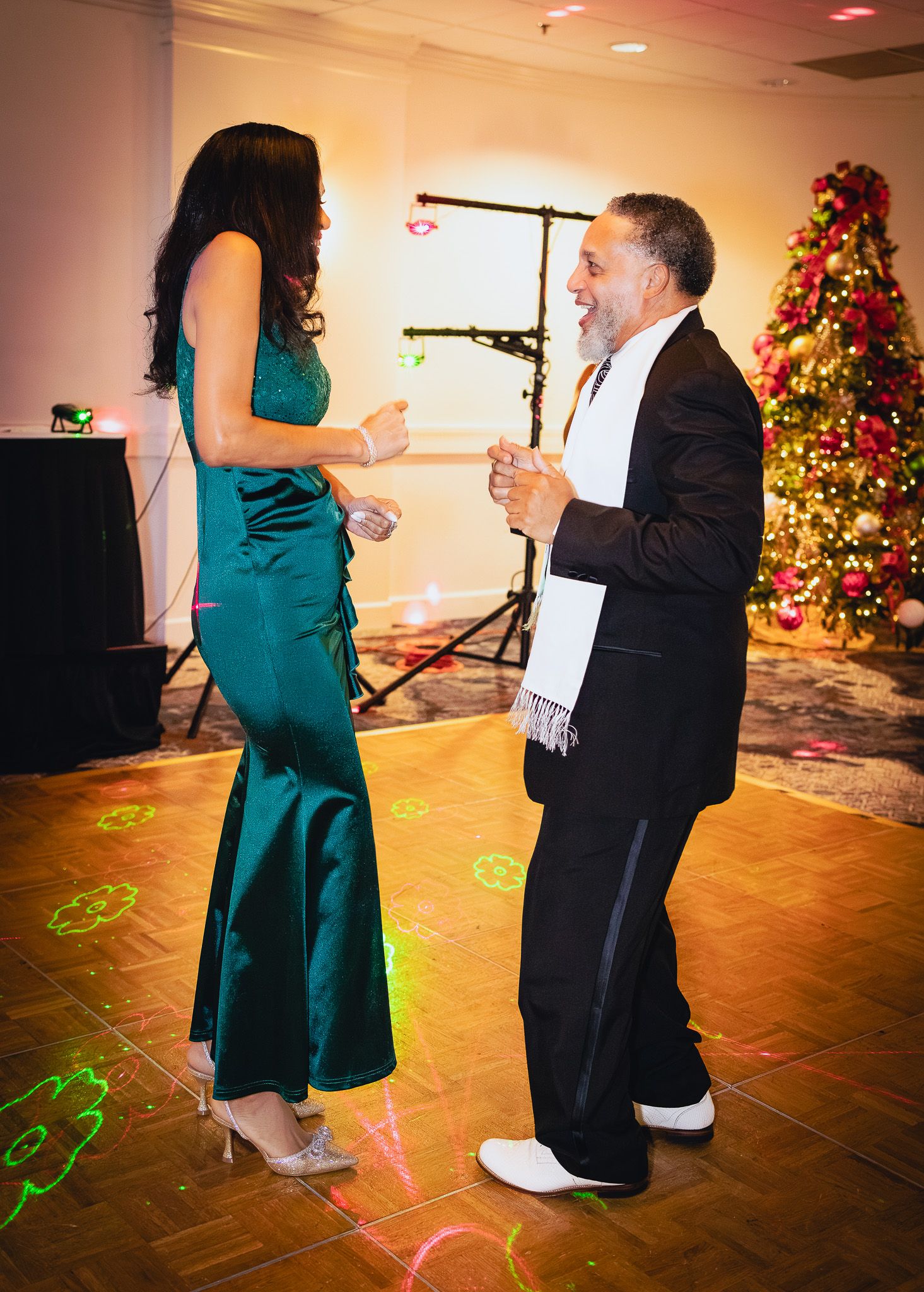 Couple dancing at a Christmas holiday party, woman in teal gown, man in black suit with white scarf, festive tree background.
