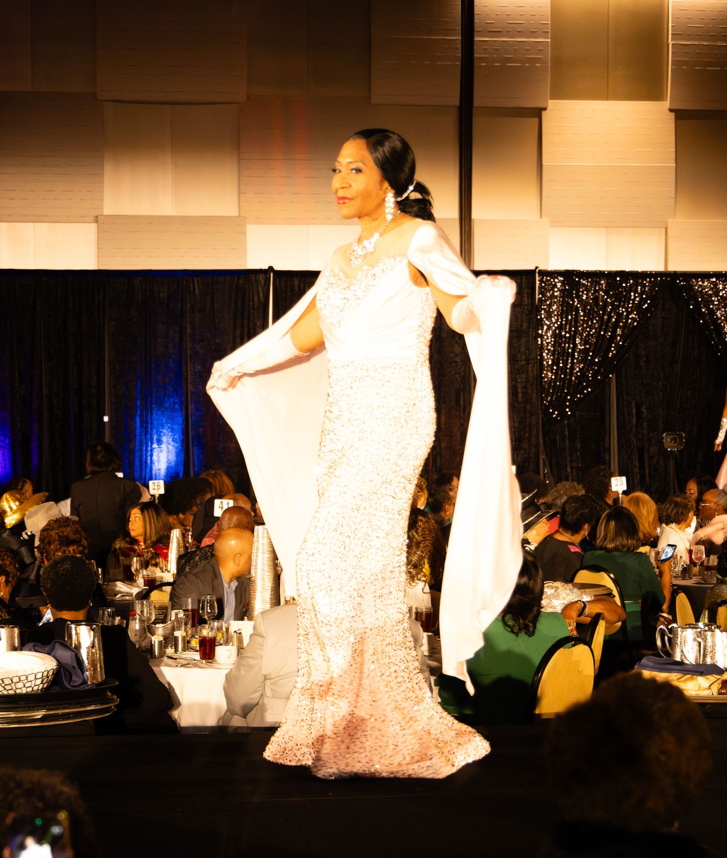 A model walks the runway in a stunning white sequined mermaid gown with a flowing cape at an elegant gala event.