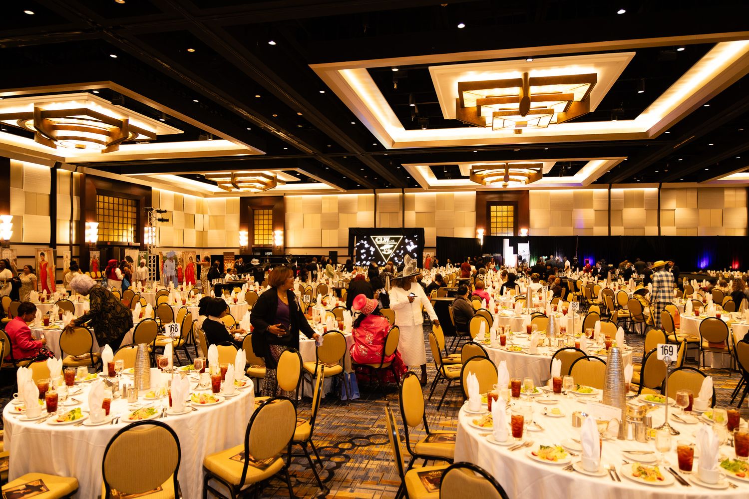 Elegant hotel ballroom banquet with round tables, gold chairs, and guests dining under decorative ceiling lights.