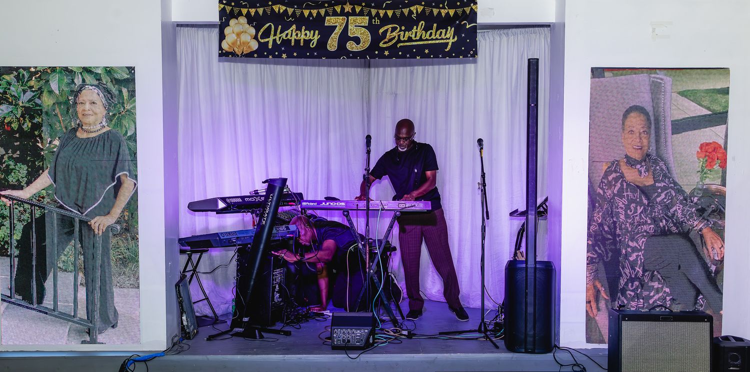 Live band performs at a 75th birthday celebration with keyboard, drums, and speakers on a decorated stage.