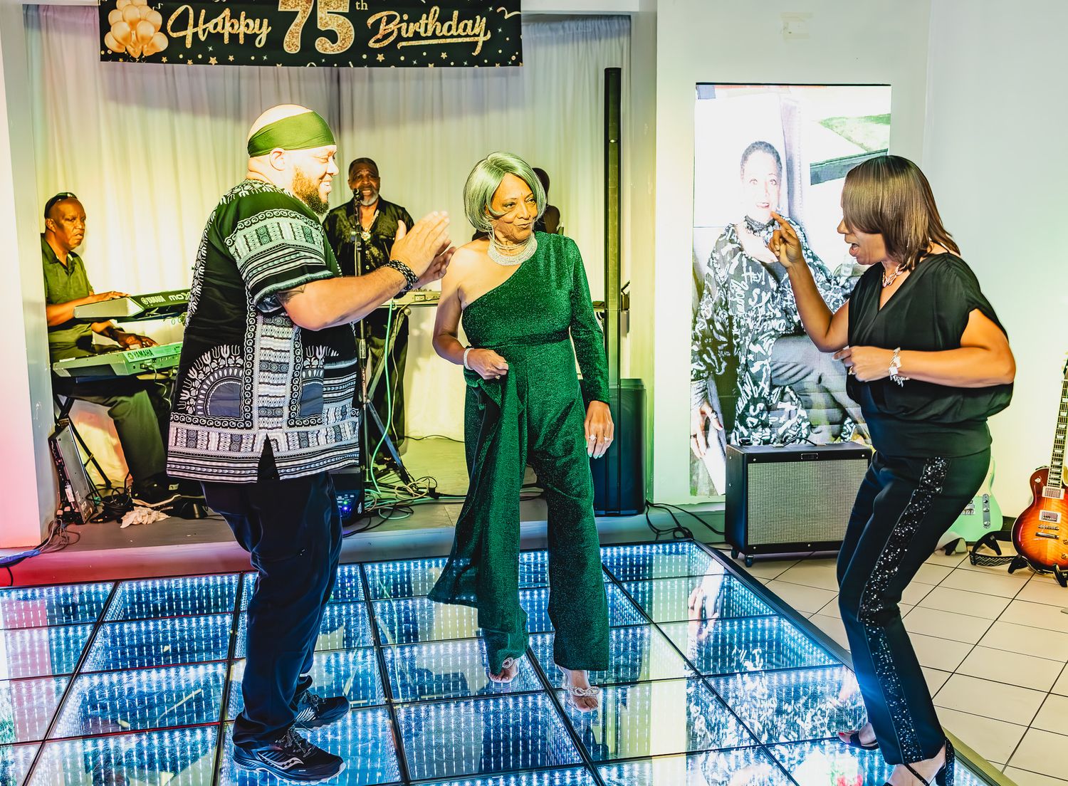 Three people dancing at a festive 75th birthday party with a live band performing in the background.