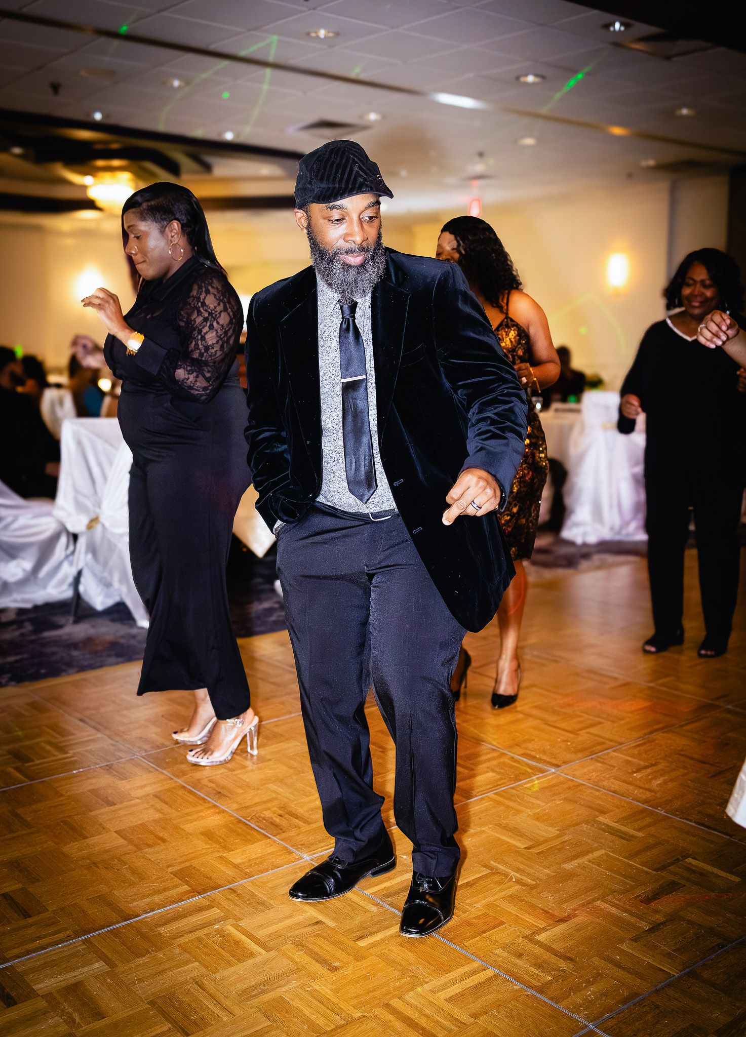 Man in velvet blazer and flat cap dancing energetically at an elegant formal event on a wooden dance floor.