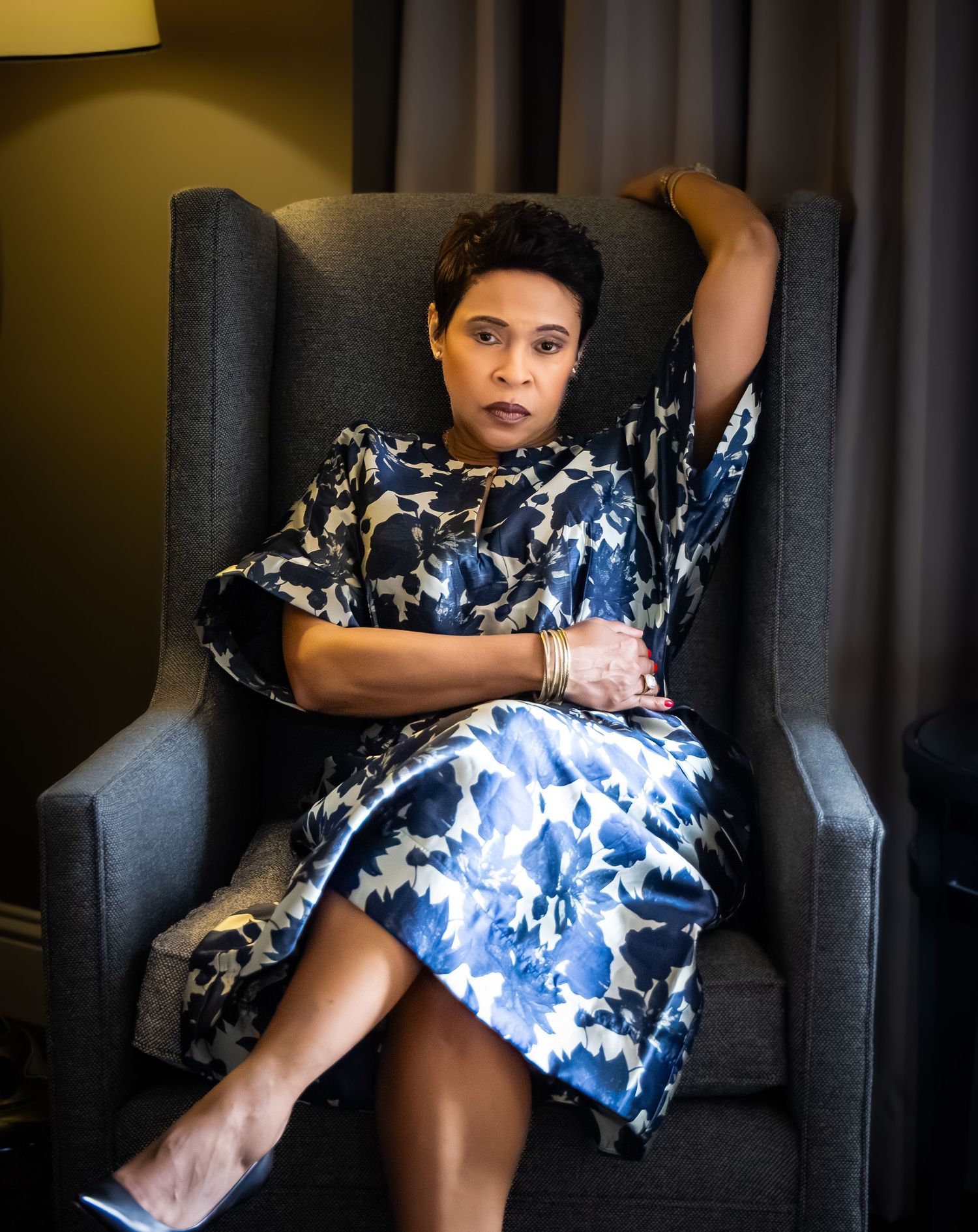 A person lounges on a dark gray armchair wearing a blue and white patterned dress in a dimly lit room.