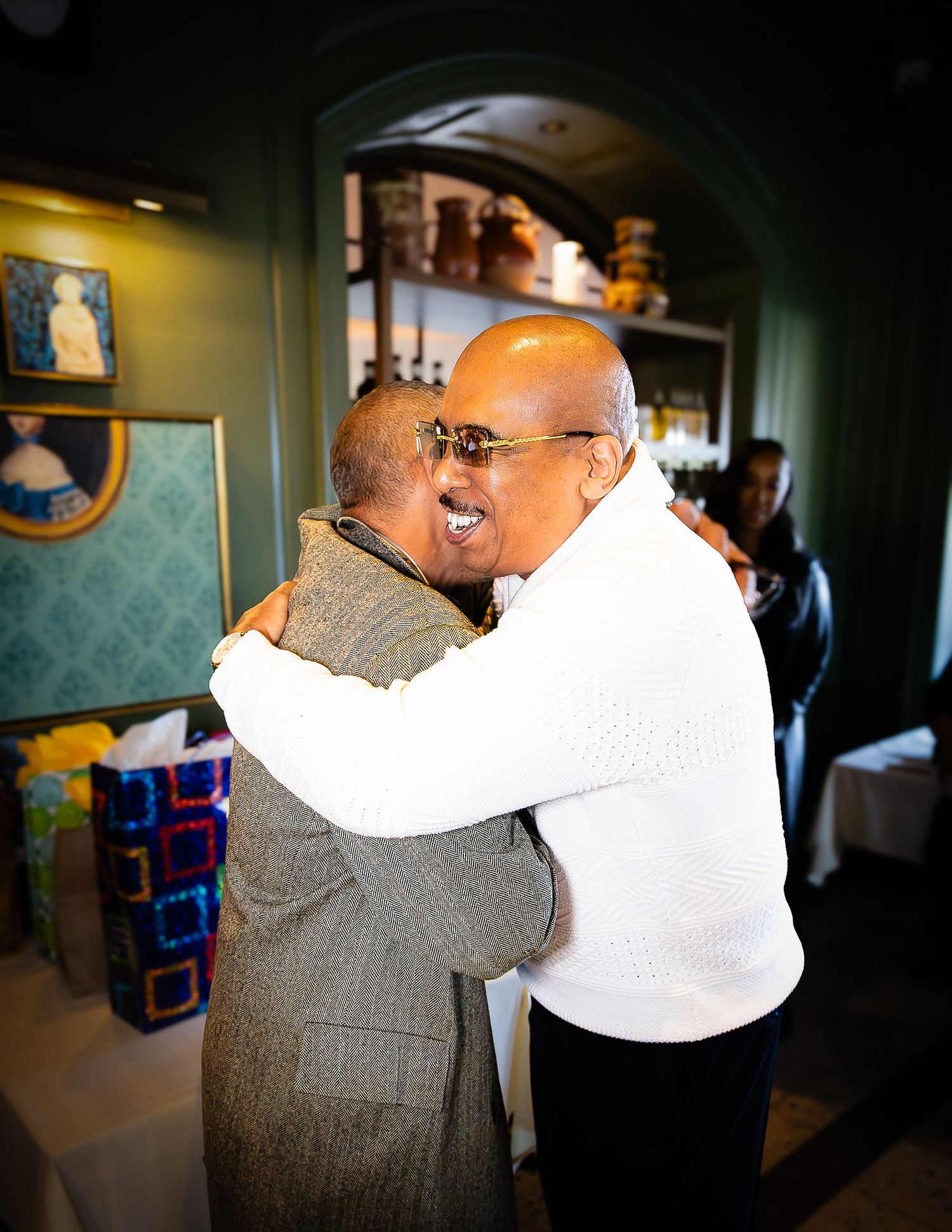 Two men share a warm embrace at an elegant restaurant gathering with green walls and decorative artwork.
