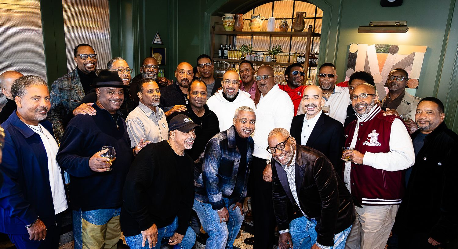 Large group of men posing together indoors, smiling for a group photo in a warmly lit room.