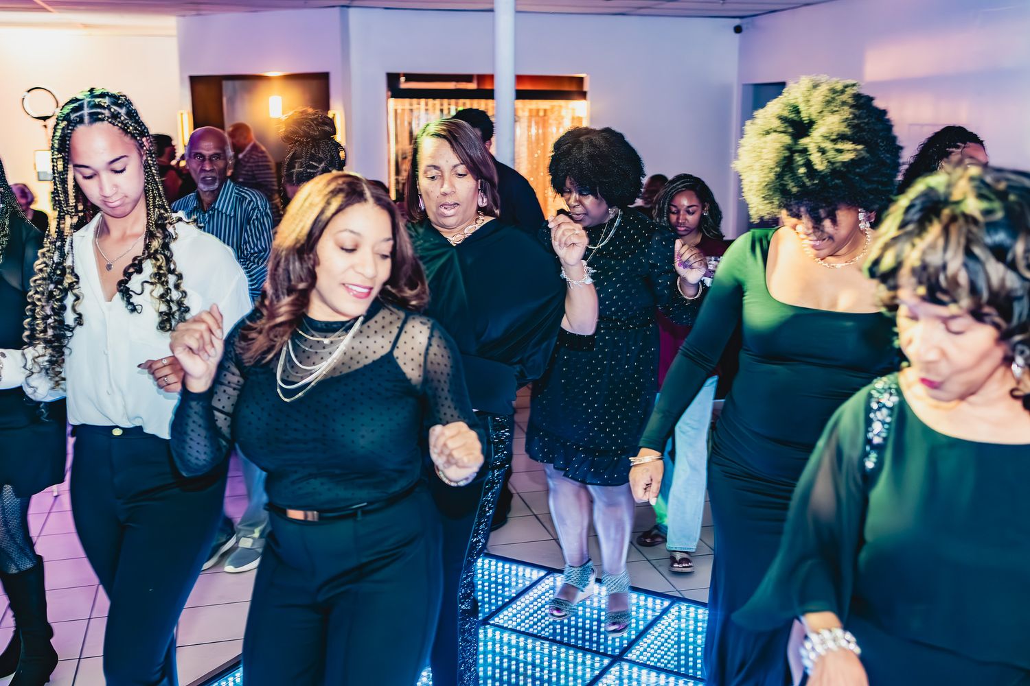 Group of women dancing energetically on a lit dance floor at a party, dressed in elegant black outfits.