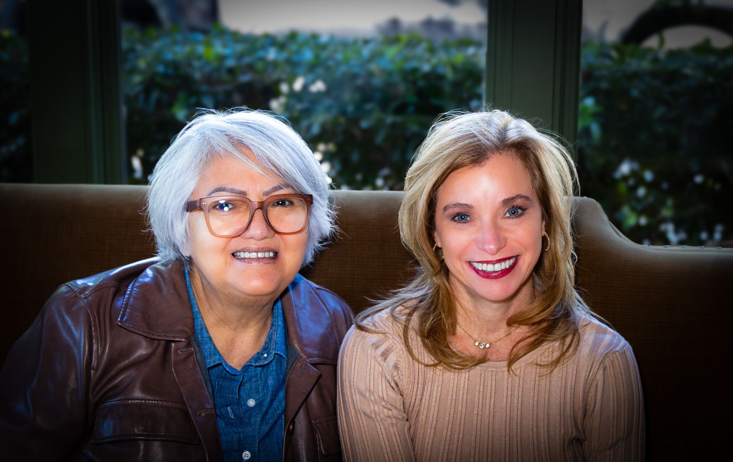 Two smiling women sitting together on a couch, one wearing glasses and a leather jacket, the other in a beige sweater.