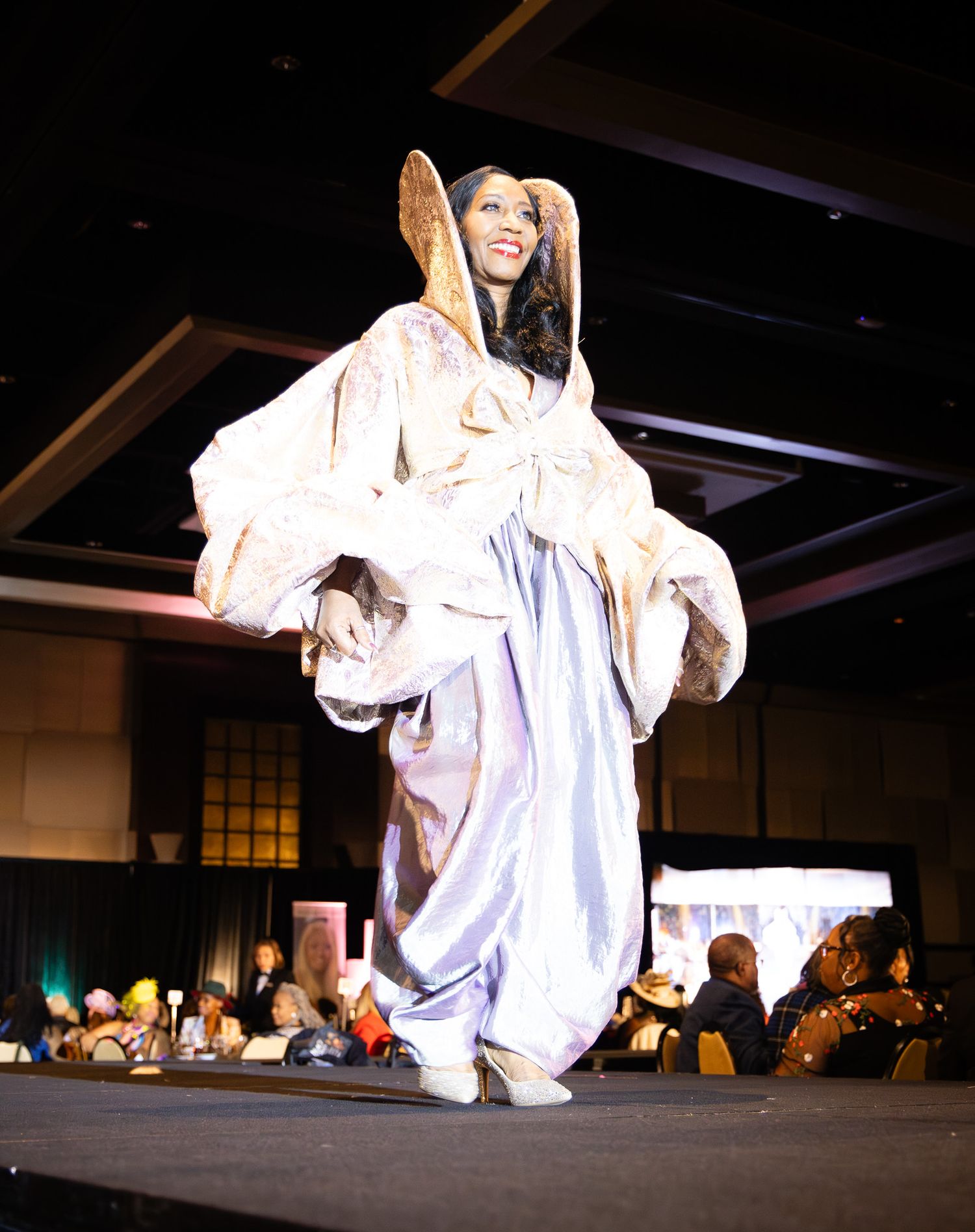 A model walks the runway in an elegant flowing lavender gown with dramatic oversized sleeves at a fashion show.