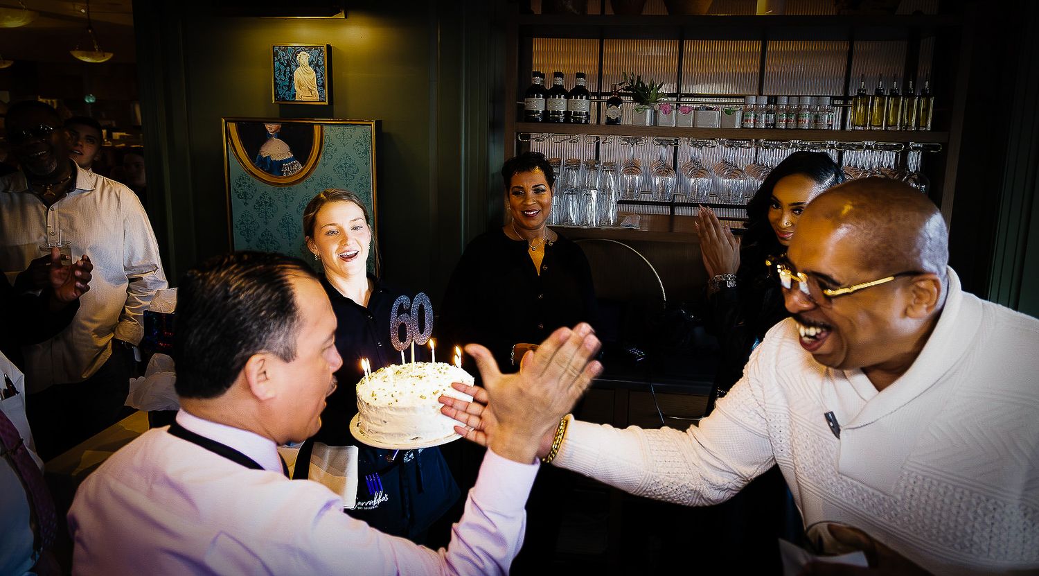 Group of people celebrating a 60th birthday with a candle-lit cake at a restaurant gathering.