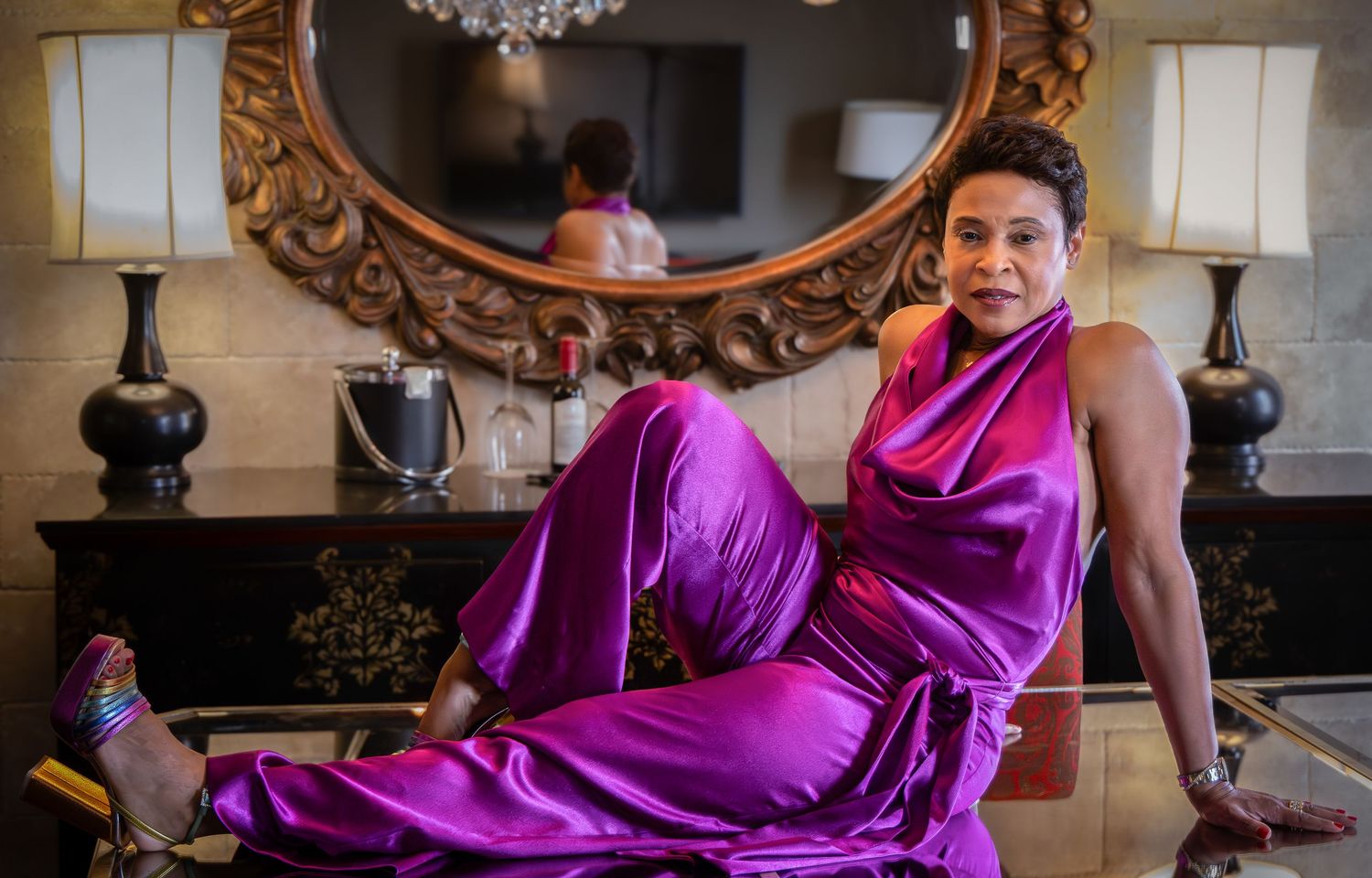 A person wearing a flowing magenta gown poses elegantly on a chaise lounge in an ornate room with chandelier.