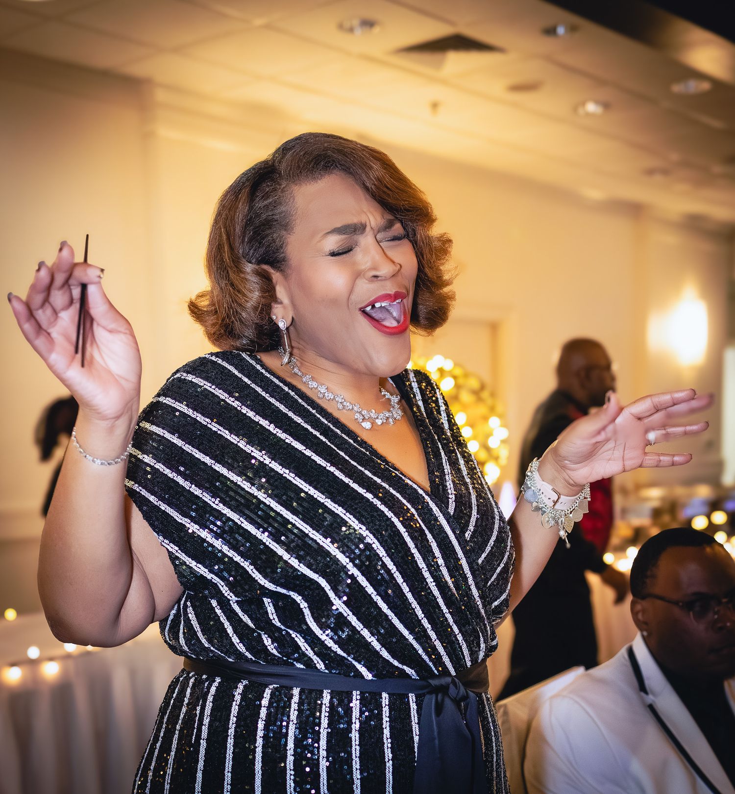 A woman in a black sequined striped dress sings passionately with hands raised at an elegant event.