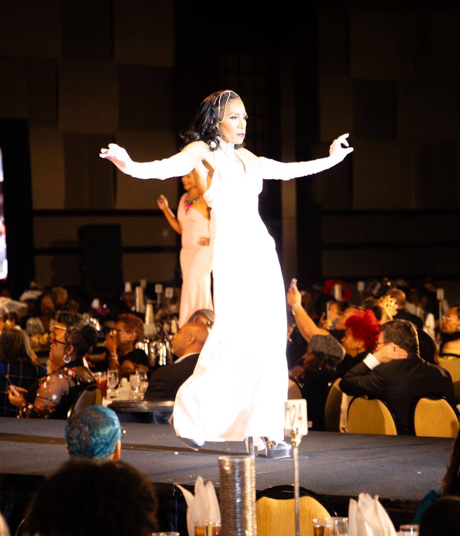 A woman in a white gown poses gracefully on a runway stage at an elegant banquet event with seated guests.