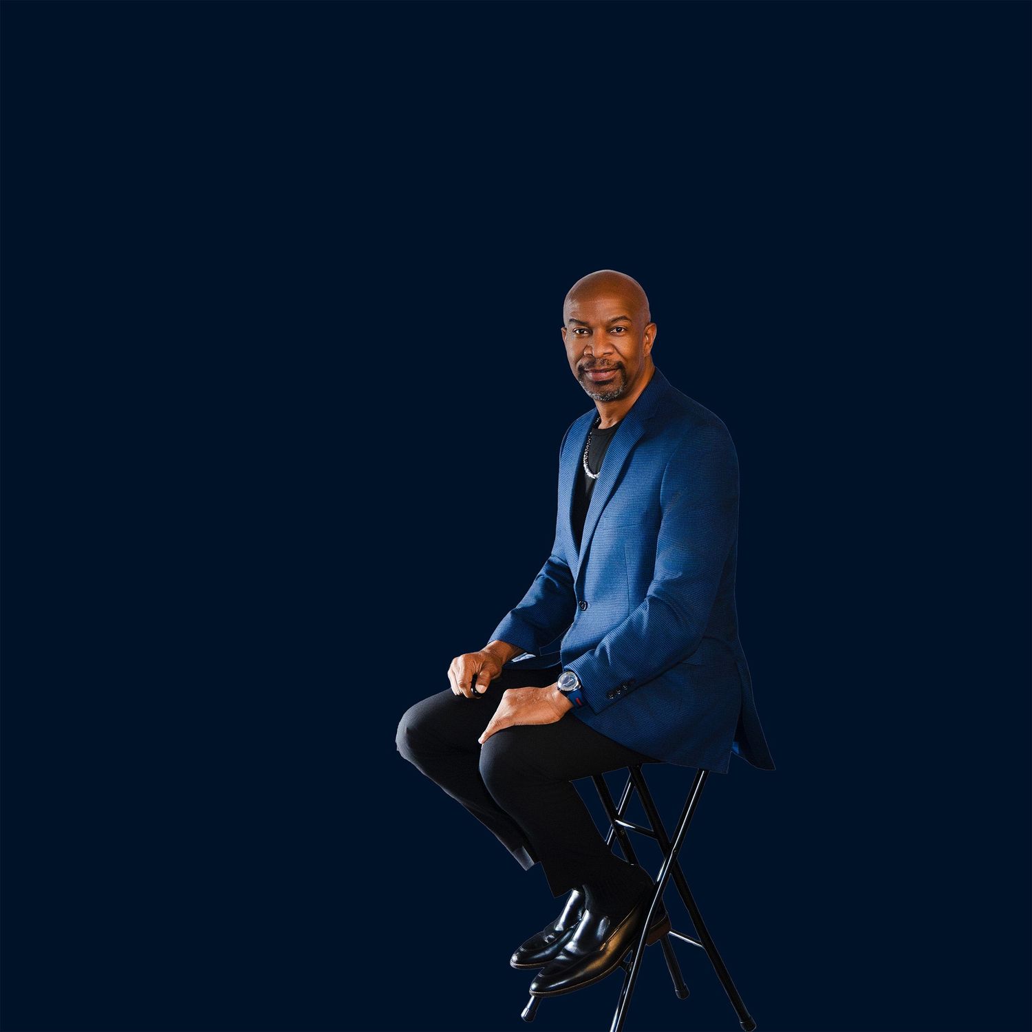 A man in a blue blazer sits confidently on a stool against a dark navy background.