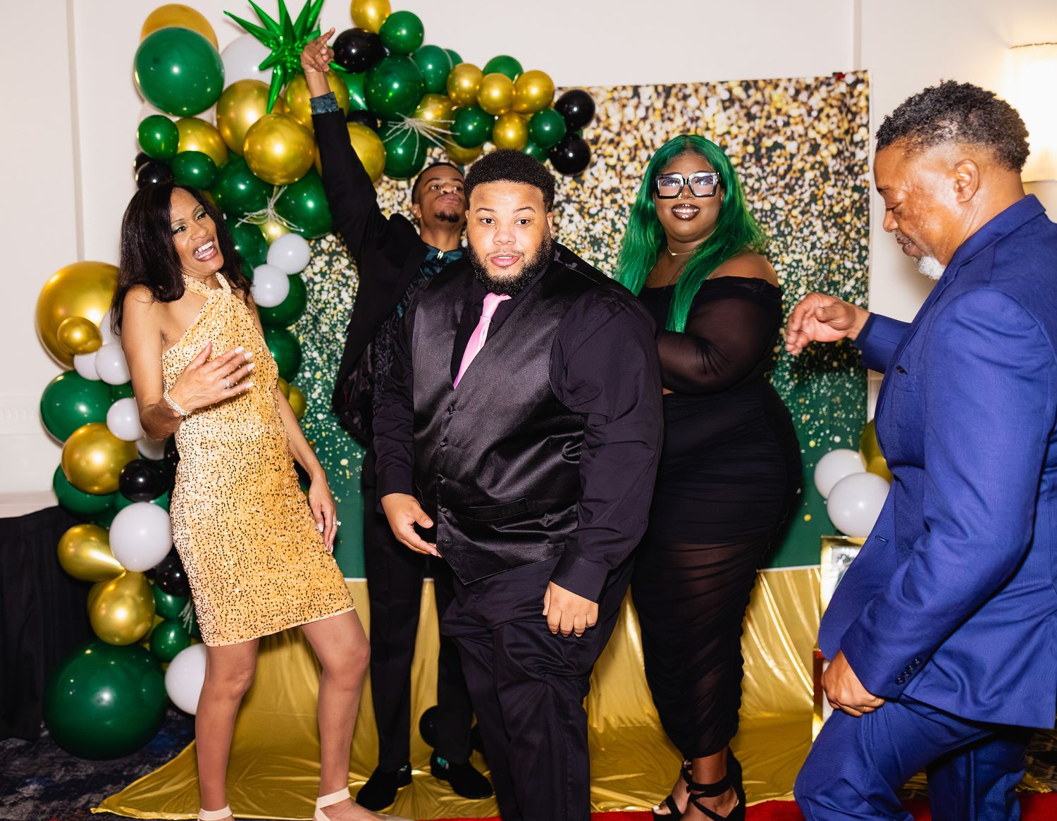 People dancing and celebrating at a festive party with green, gold, and white balloon decorations.