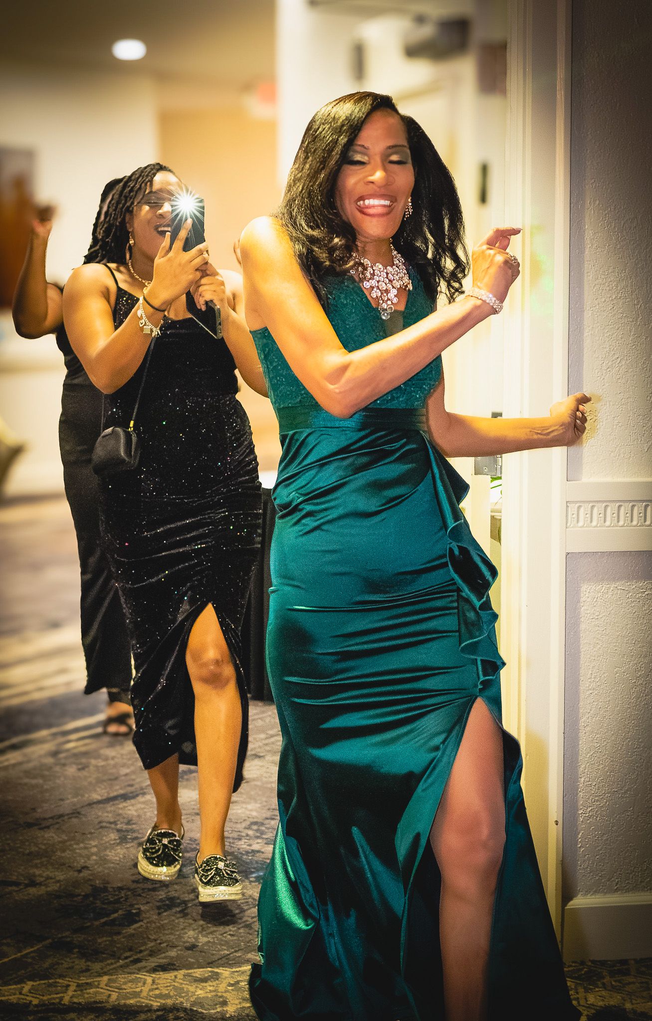 Woman in elegant teal green gown poses in hallway while friend photographs her at formal event.