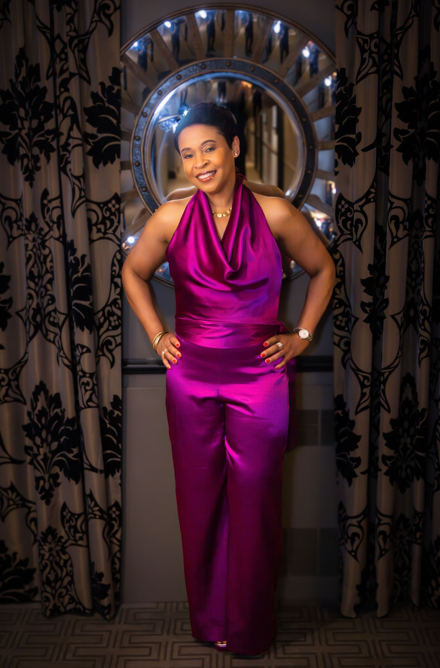Elegant figure in a vibrant magenta satin jumpsuit poses confidently against an ornate archway with decorative lighting.