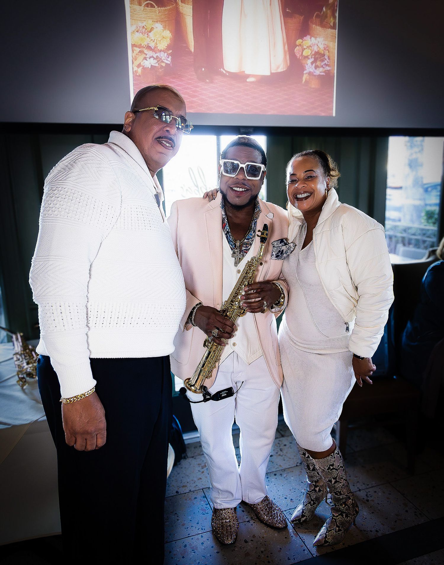 Three people in white outfits posing together, with center person holding a saxophone at an elegant indoor event.