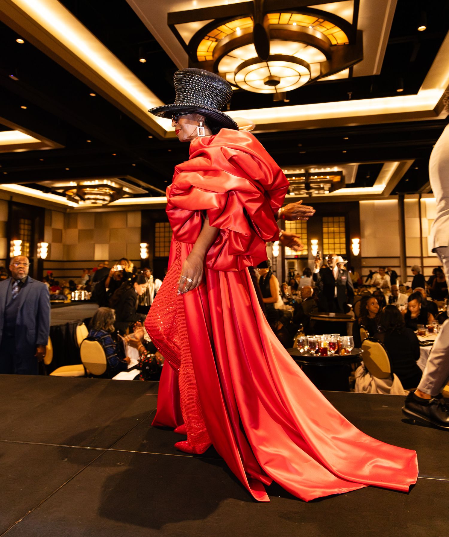 Model walks runway in dramatic red satin gown with ruffled shoulders and wide-brim black hat at elegant ballroom fashion show.