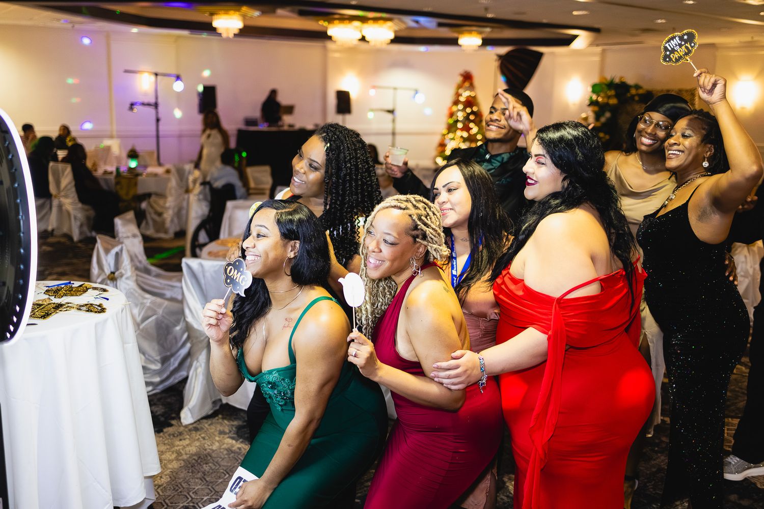 Group of women in elegant dresses posing and dancing at a festive holiday party with Christmas tree in background.