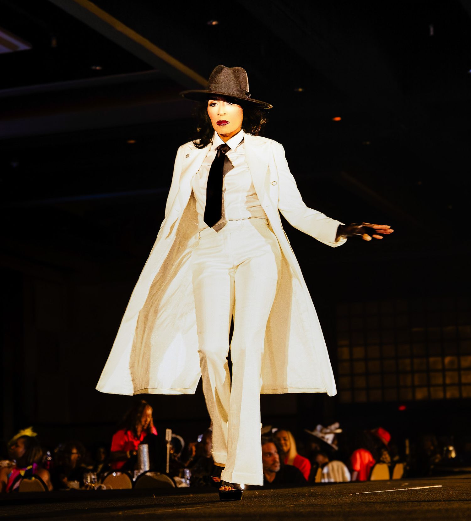 Model walks runway in white suit, long white coat, black tie and fedora hat during a fashion show.