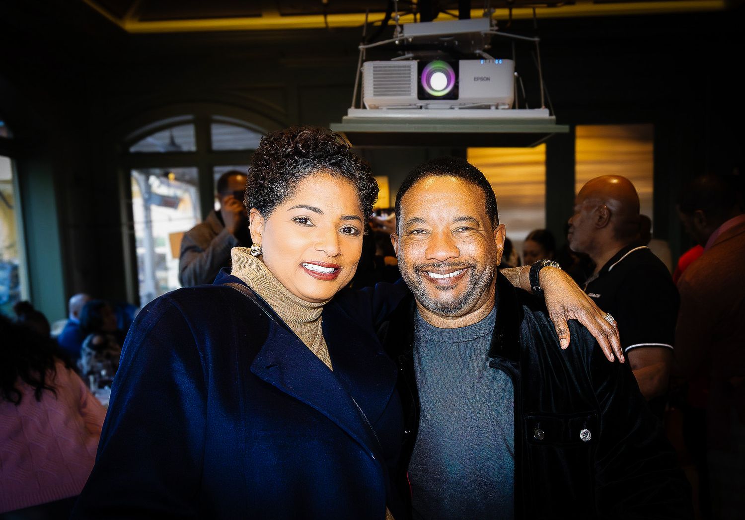 Two smiling people pose together at an indoor social event with a projector visible in the background.