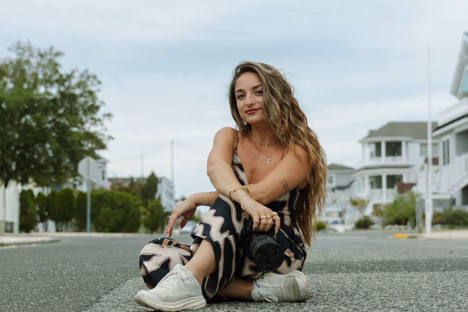 Young woman with long wavy hair sitting on a street road, wearing patterned pants and white sneakers, smiling outdoors.