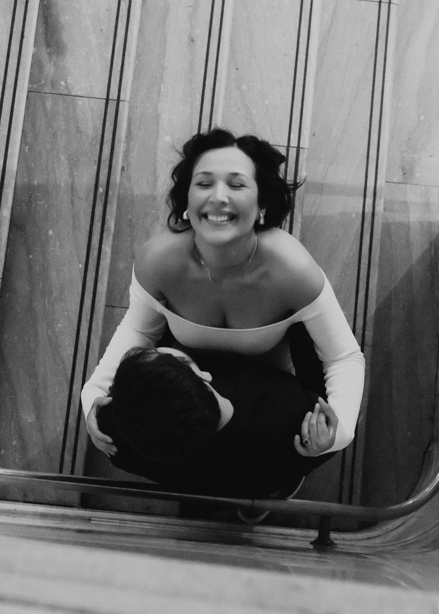 Black and white photo of someone crouching in an off-shoulder white top against geometric lines.