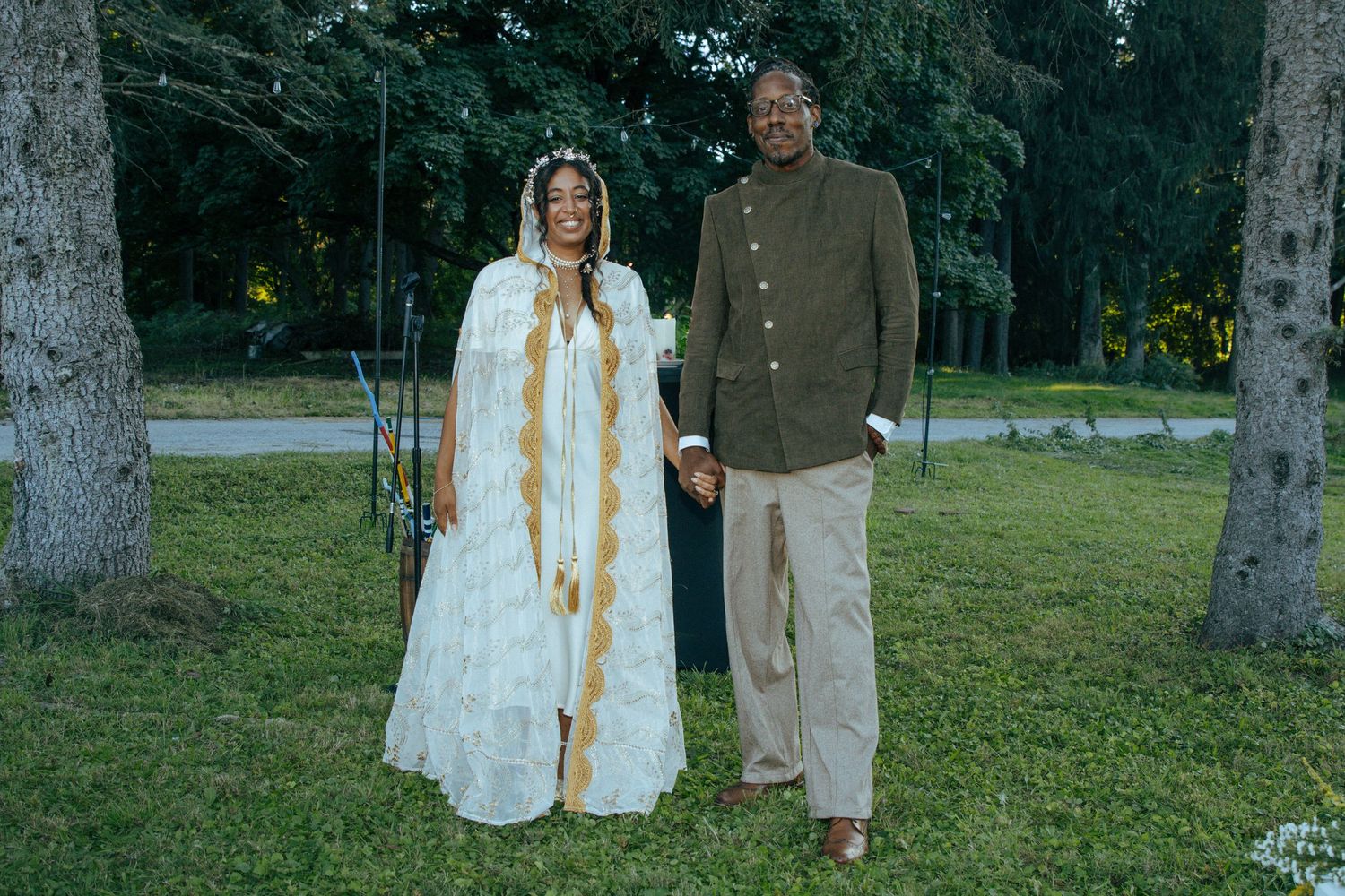 Couple in white and earth-toned attire standing among trees in a park setting.