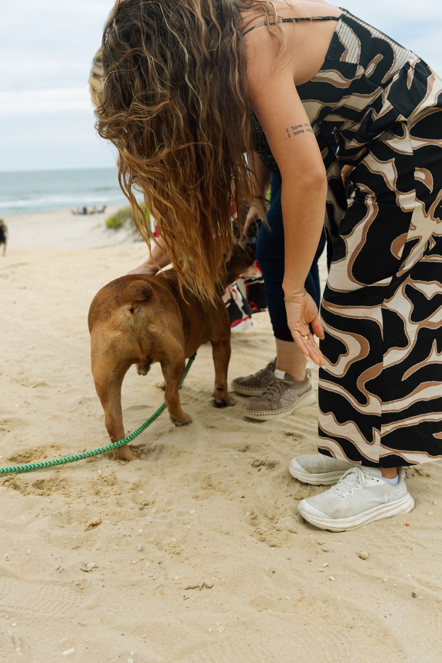 Woman in printed dress bending down to pet a brown dog on a sandy beach.