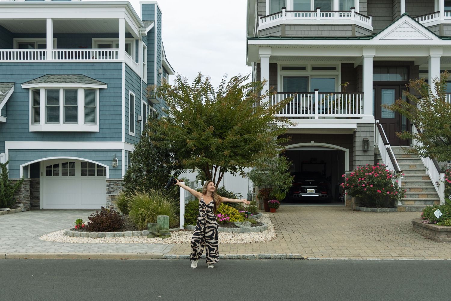 A woman in a patterned outfit stands on a brick driveway between two large coastal homes with porches.