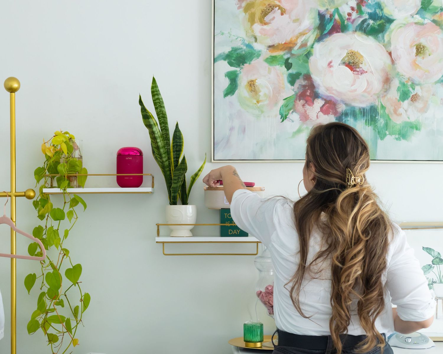 Woman arranging plants and decor on gold wall shelves beside a floral painting in a bright, modern living space.