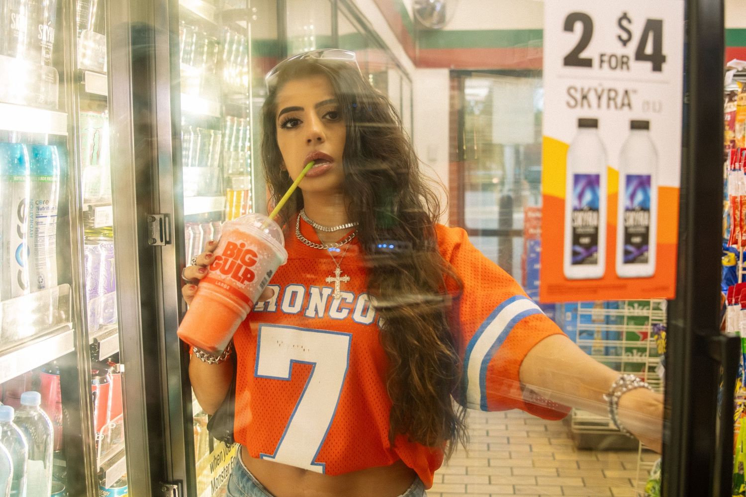 Young woman in orange Broncos jersey holding a drink inside a convenience store with refrigerated shelves.