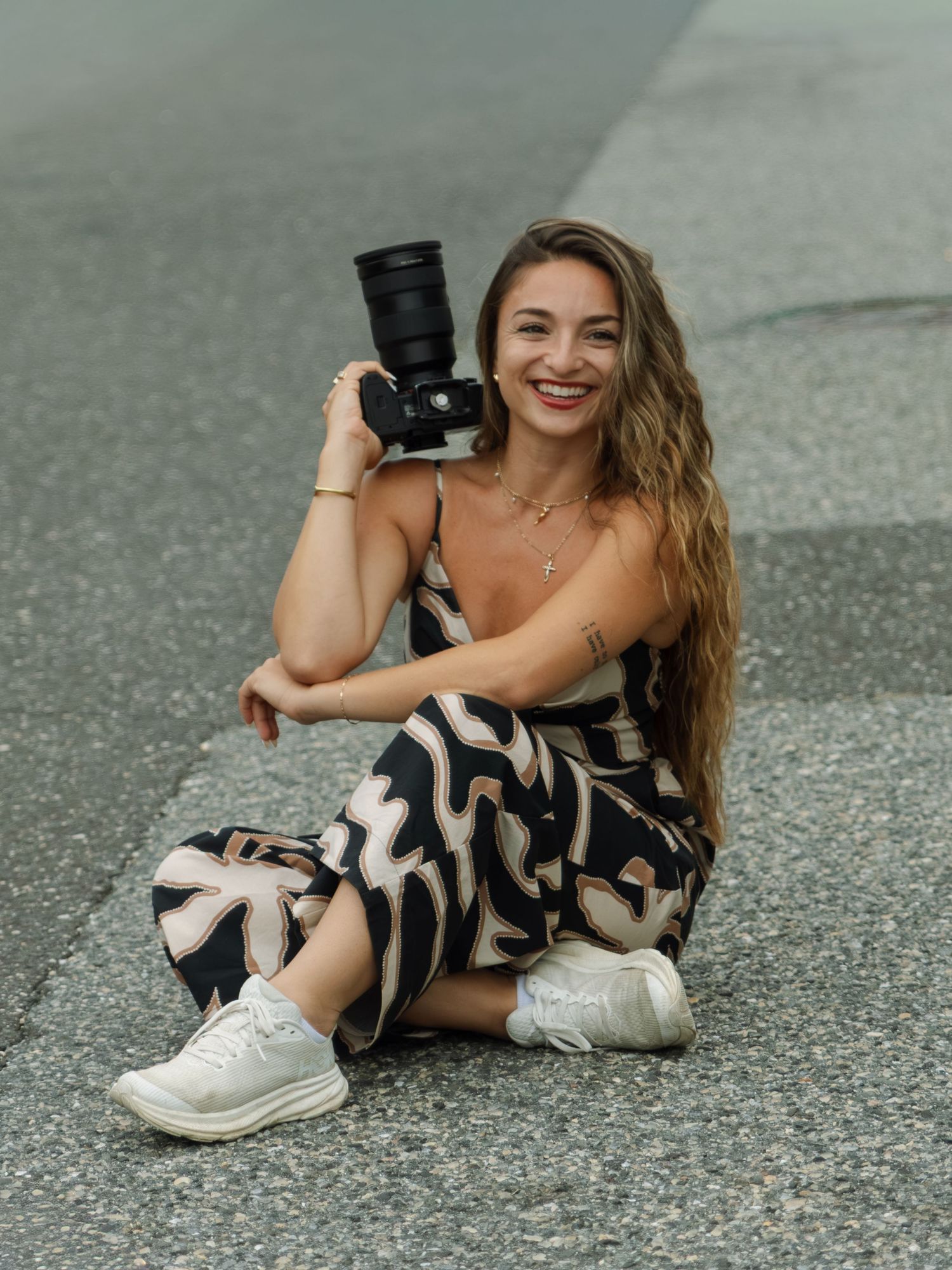 Smiling woman sitting on pavement holding a camera, wearing a floral jumpsuit and white sneakers.