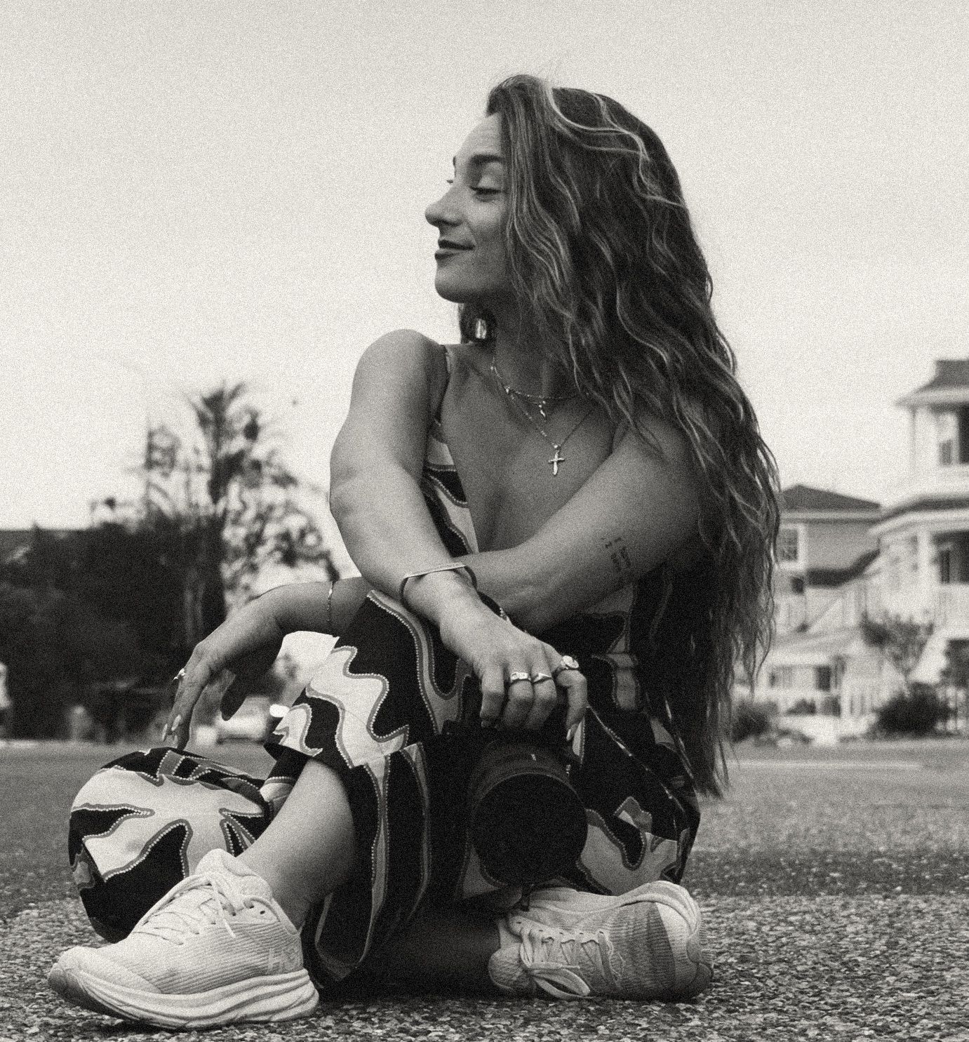 Black and white photo of a young woman with long wavy hair sitting outdoors, holding a camera, wearing sneakers.
