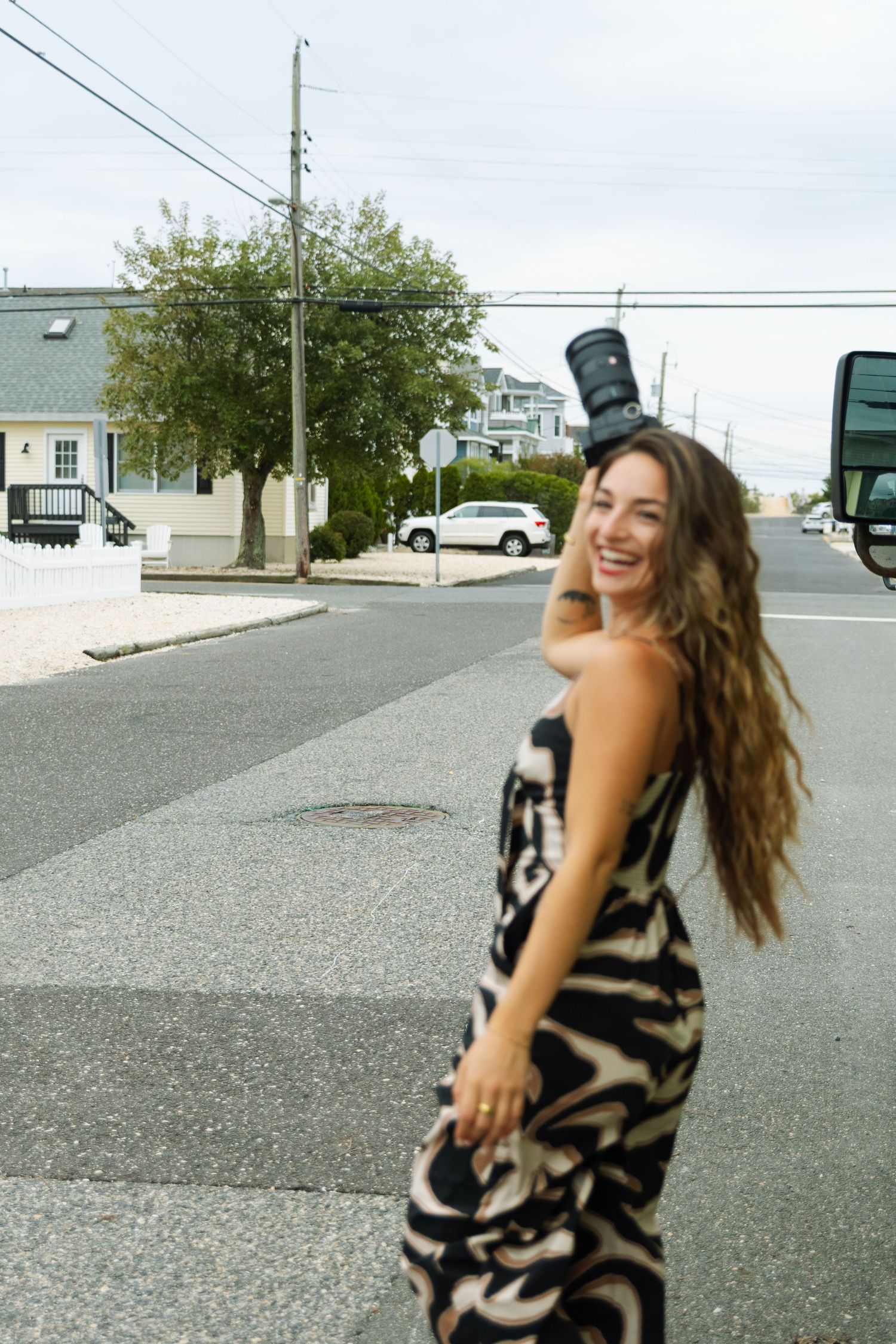 Smiling woman in patterned dress balancing camera on head while crossing a street in a coastal town.