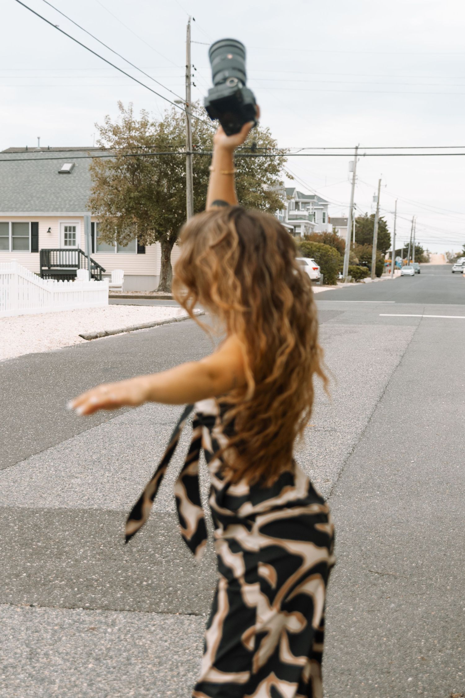 Woman in patterned jumpsuit holding camera overhead while posing on a suburban street.