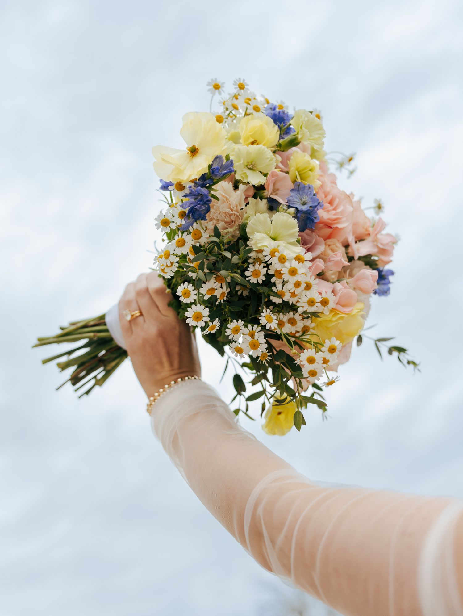 Hand holding a vibrant wildflower bouquet with daisies and colorful blooms against a light background.