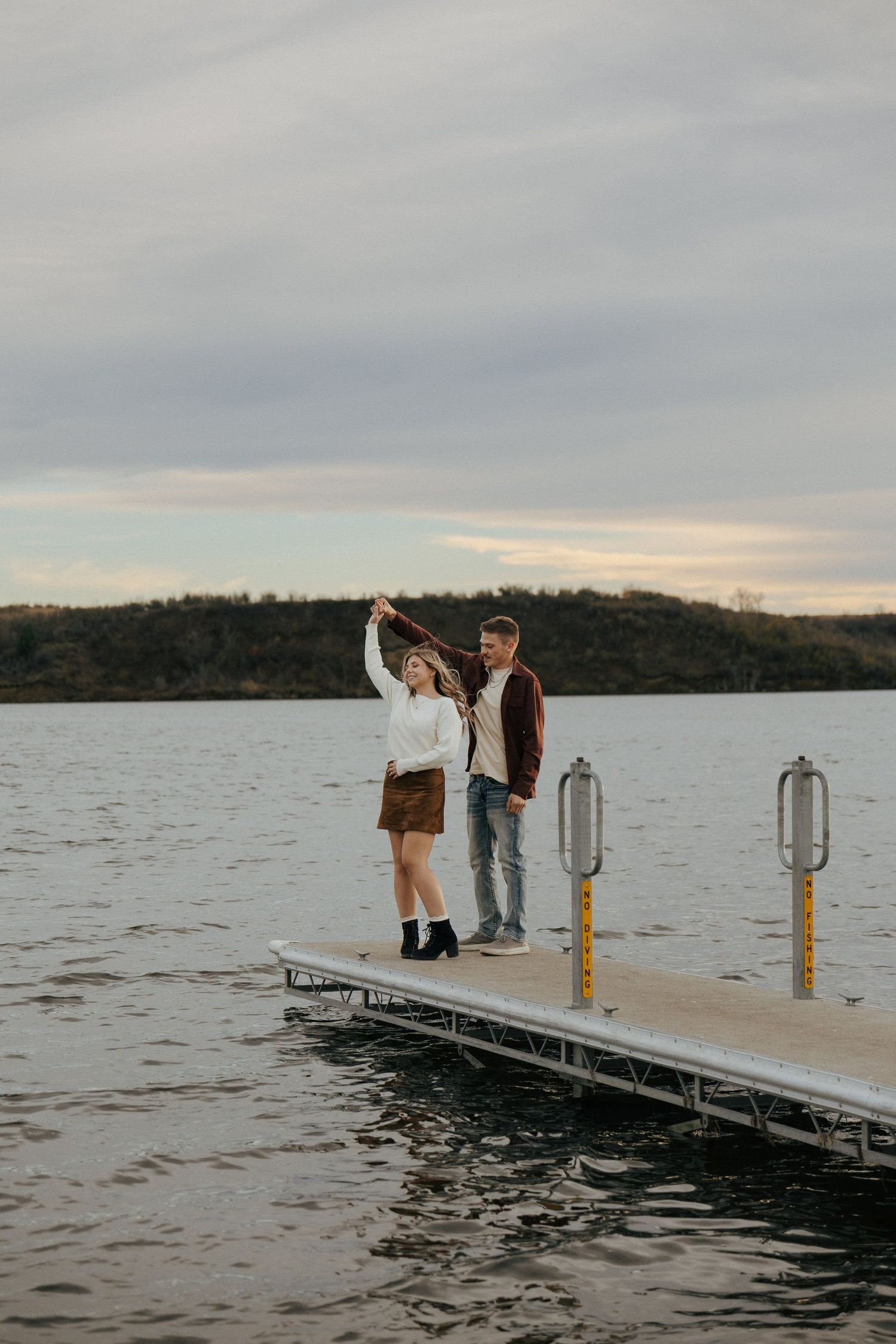 Two people stand at the end of a dock on a lake during sunset.