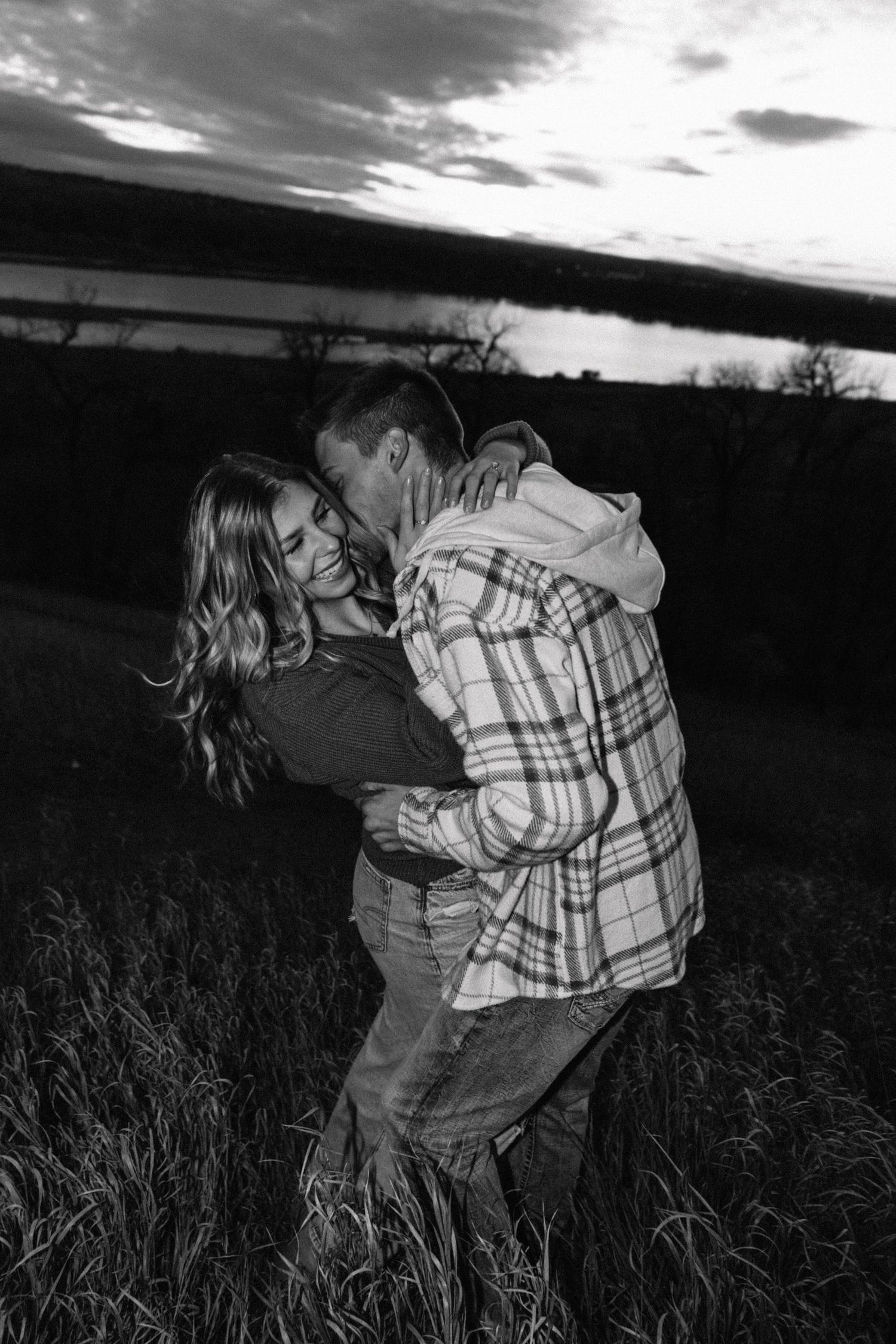 Romantic couple embraces in a field at sunset with dramatic clouds in the background.