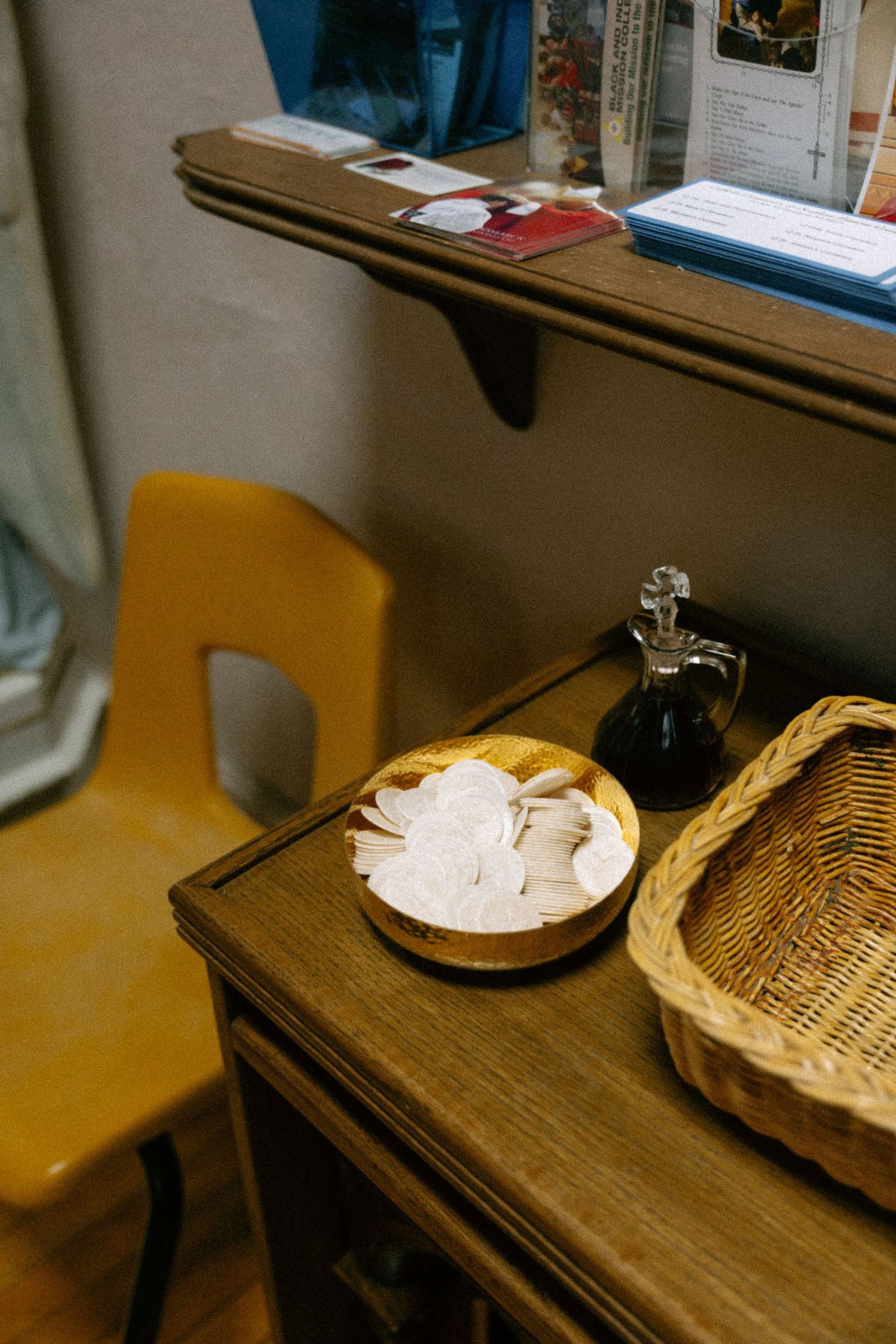 Wooden desk with communion wafers in bowl, oil bottle, wicker basket, yellow chair, and shelf with books in church setting.