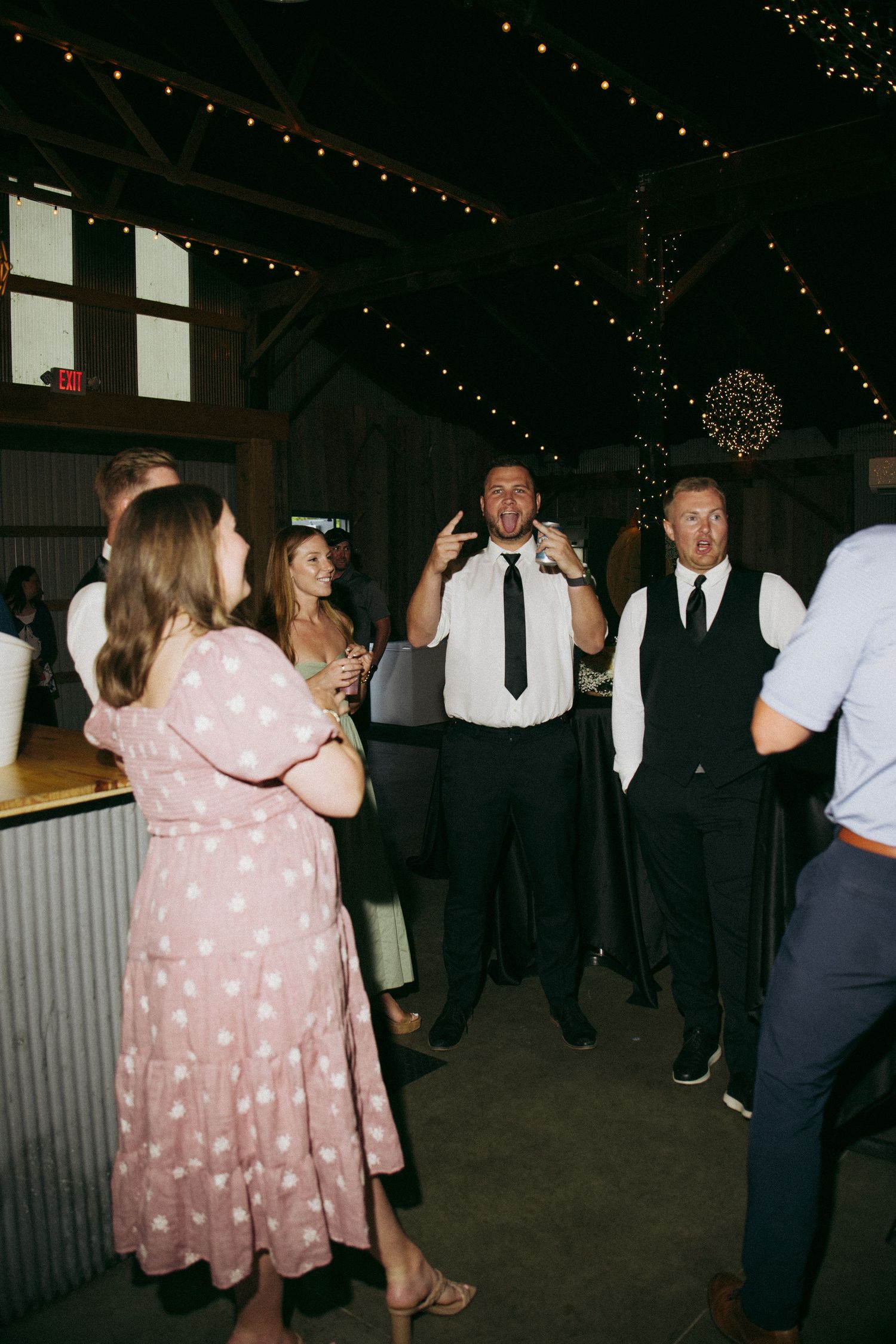 Wedding guests dancing and celebrating inside a rustic barn venue decorated with string lights at night.