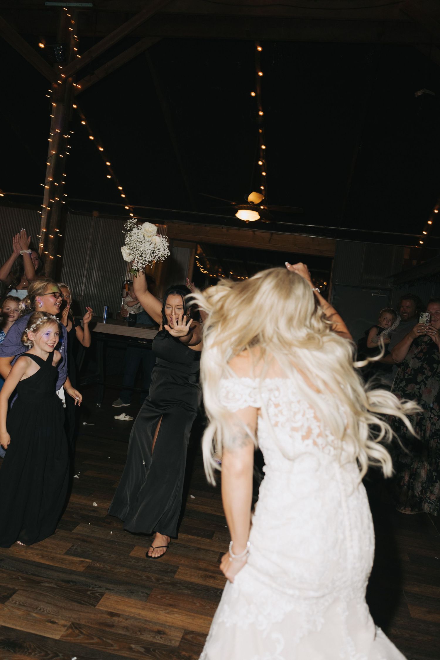 Bride in lace mermaid gown tosses bouquet to excited guests on the dance floor at rustic barn reception.