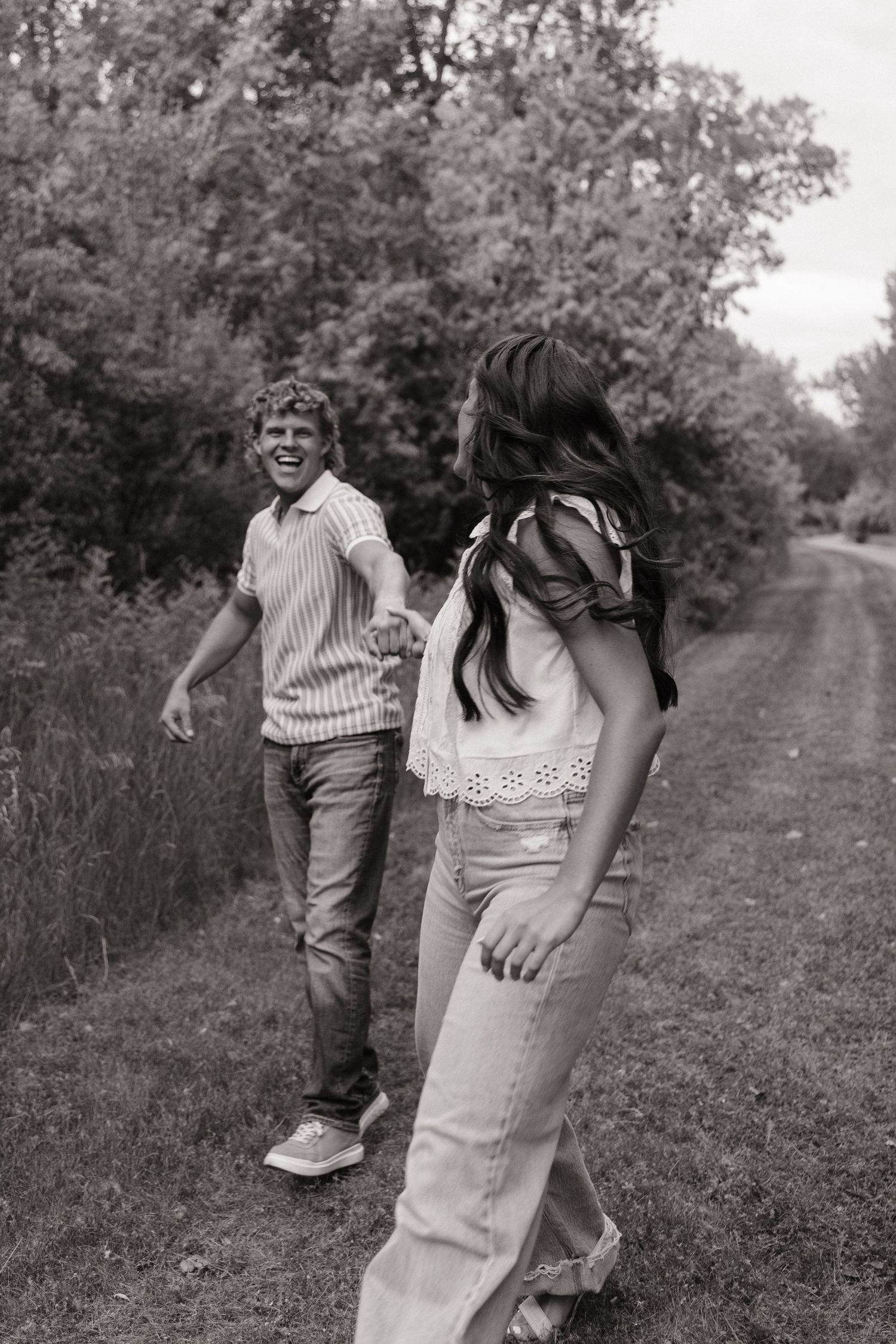 Black and white photo of a laughing couple holding hands while walking along a scenic rural path surrounded by lush trees.