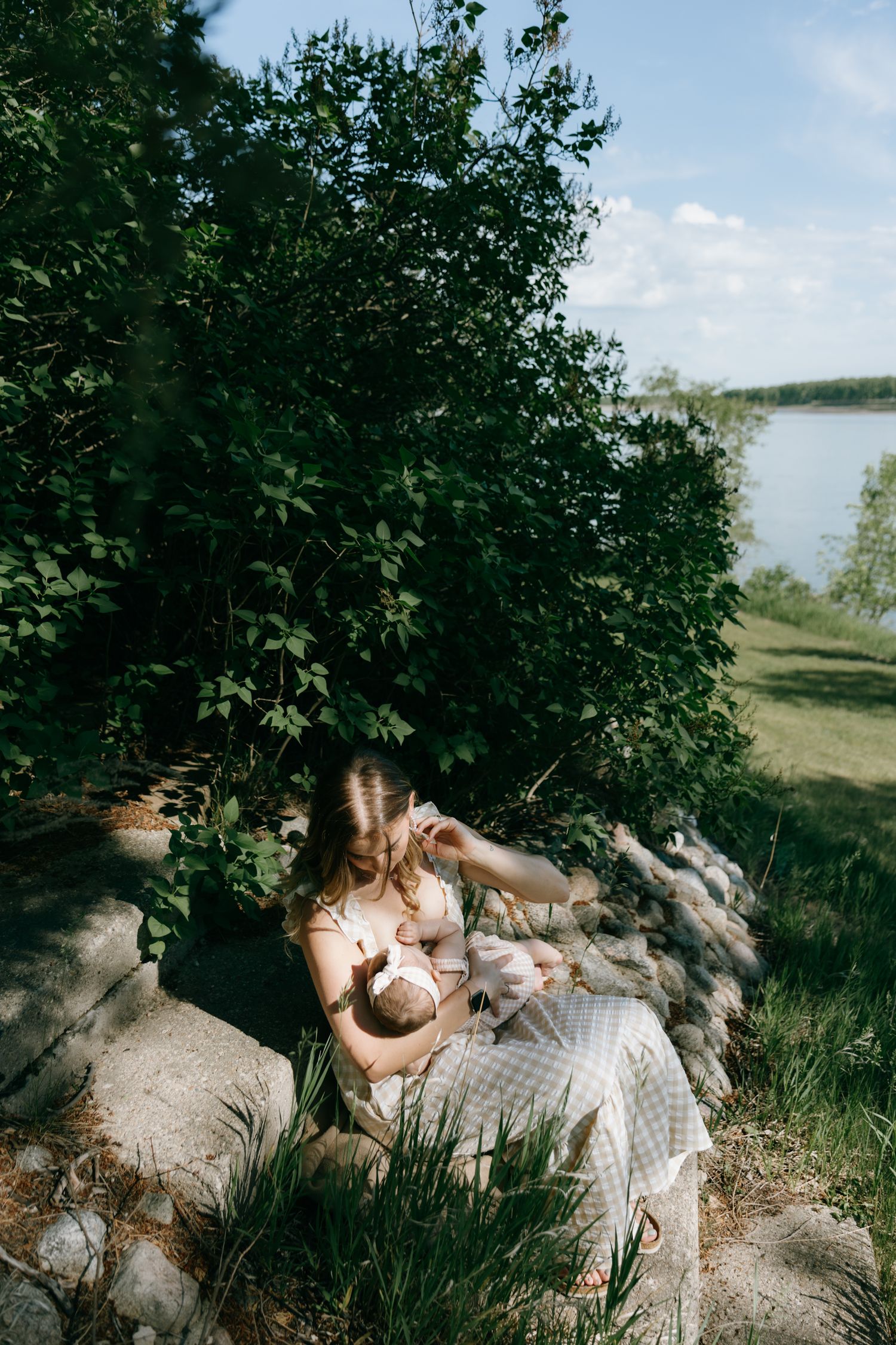 Mother breastfeeding newborn baby outdoors near lake, sitting on rocks surrounded by lush green trees.